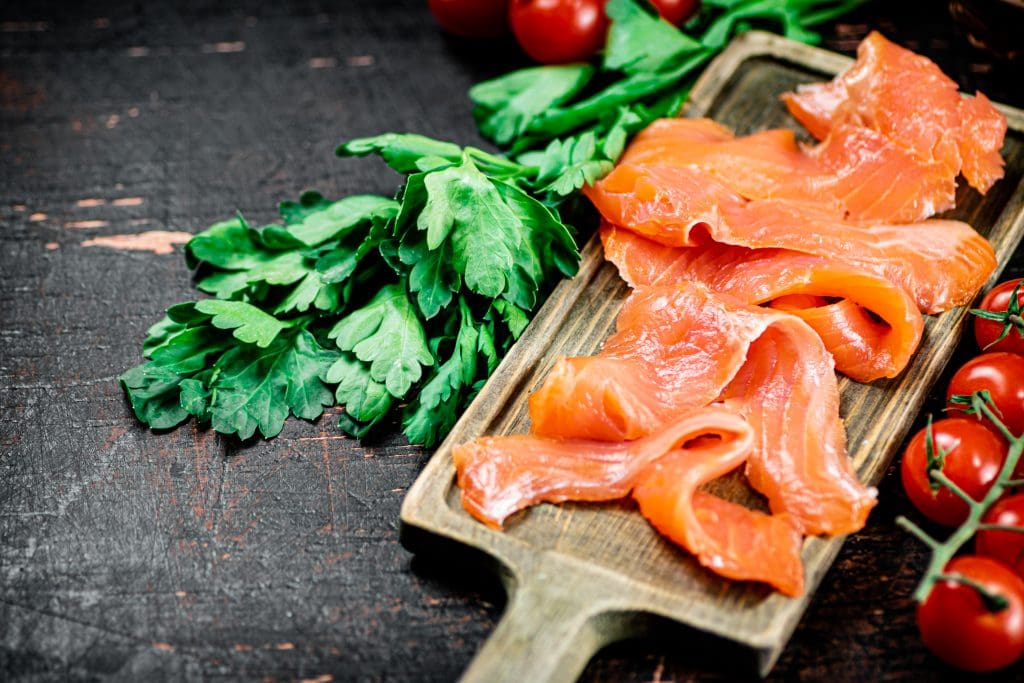 Slices of salted salmon with greens and tomatoes on a cutting board.