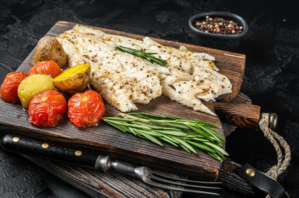 Baked Haddock fish fillet on wooden board with tomato and potato. Black background. Top view