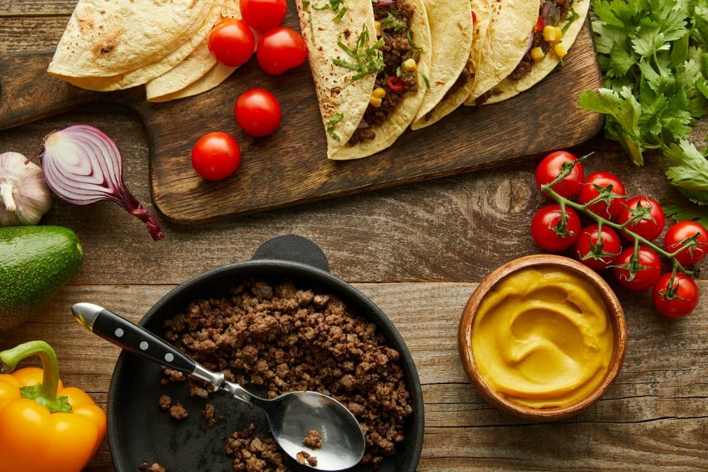 Top view of delicious tacos with minced meat on cutting board and ingredients on wooden background